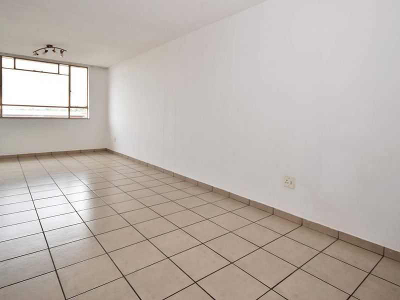 A bright, empty living room featuring neutral tiled flooring, a large window providing natural light, and standard electrical fixtures.