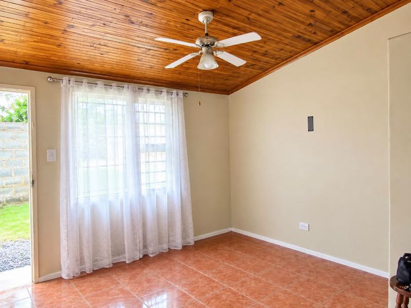 A bright, empty living room featuring a timber-clad ceiling, ceiling fan, tiled flooring, and direct access to the garden through a security door.