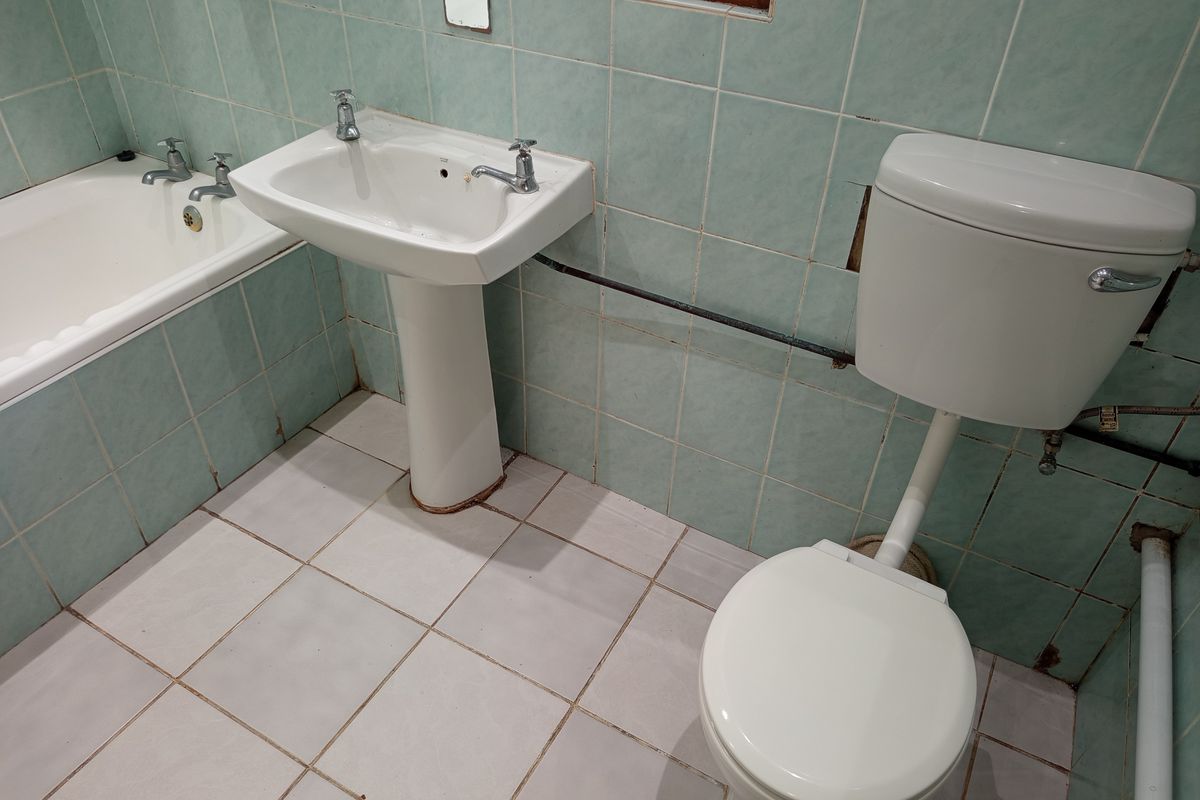 A dated bathroom featuring a pedestal basin, bathtub, and toilet with original light blue wall tiling and exposed plumbing fixtures.