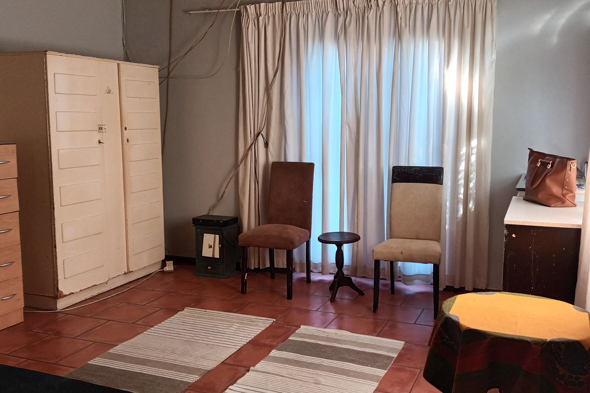 Living area featuring terracotta floor tiles and large window with curtain tracks. The space requires cosmetic updates and electrical cable management.