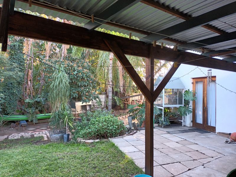 A covered outdoor patio area featuring timber support beams, a corrugated metal roof, and paved flooring, overlooking a garden space.