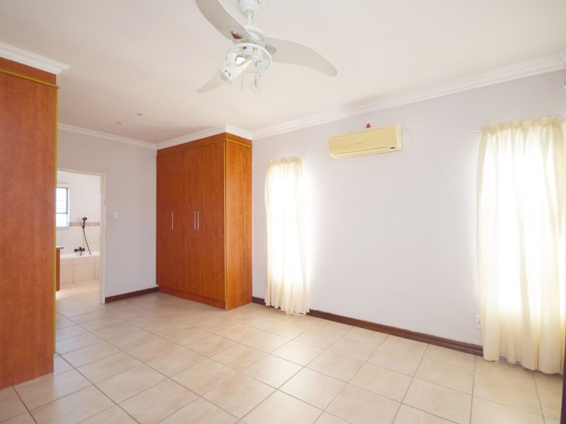 Spacious bedroom featuring tiled flooring, built-in cupboards, a ceiling fan, and a wall-mounted air conditioning unit, with an adjoining en-suite bathroom.