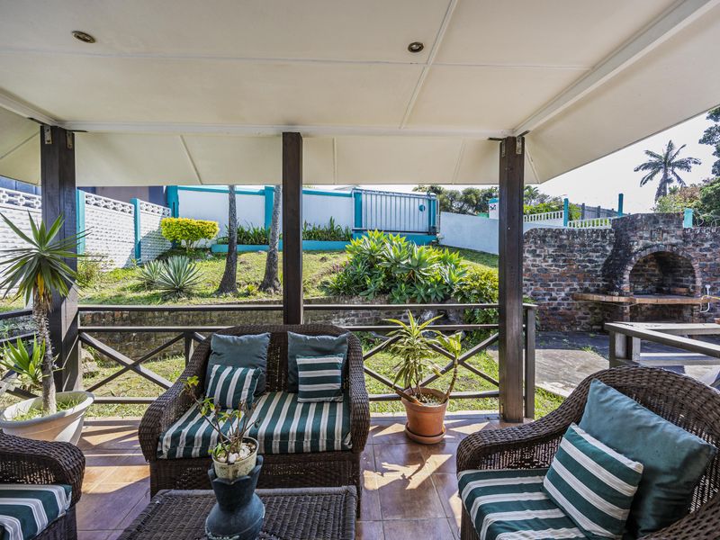 A covered outdoor patio featuring tiled flooring, a built-in brick braai, and a timber balustrade overlooking a landscaped garden area.