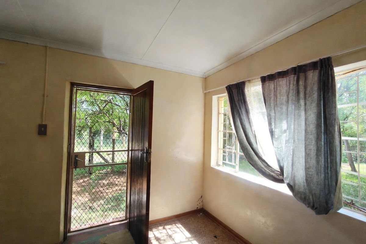 An empty bedroom featuring a security gate, timber door, and steel-framed windows, requiring cosmetic updates to the walls and flooring.