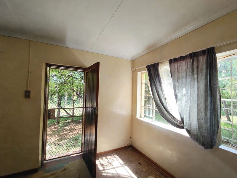 An empty bedroom featuring a security gate, timber door, and steel-framed windows, requiring cosmetic updates to the walls and flooring.