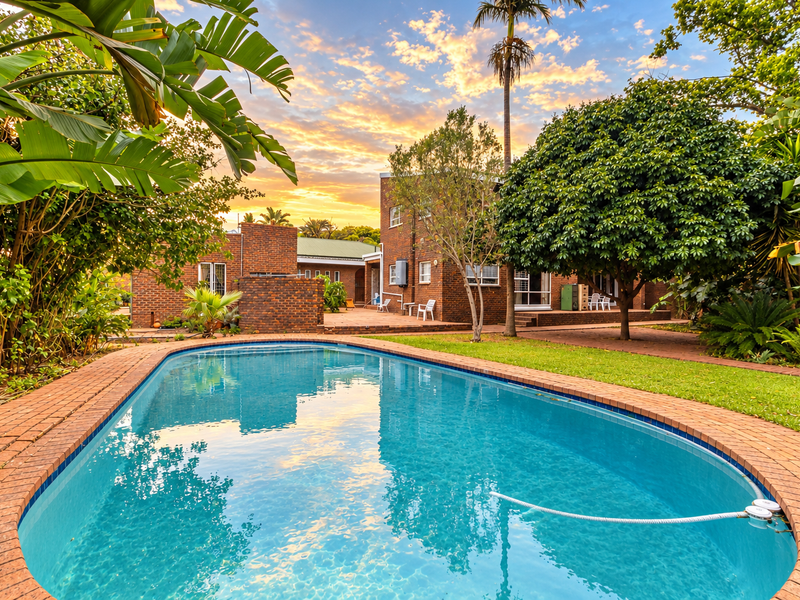A well-maintained swimming pool set within a lush garden, featuring brick paving and a view of the rear brick facade of the house.