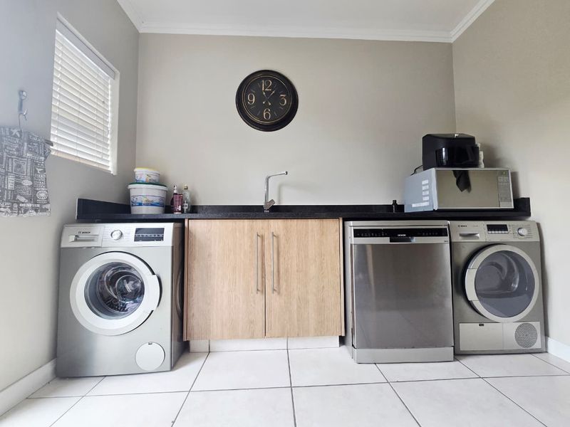A functional laundry area featuring built-in cabinetry, a sink, and space for multiple appliances, finished with tiled flooring and neutral wall tones.