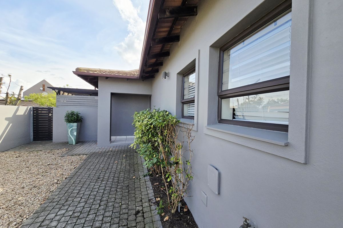 A well-maintained rear exterior view featuring a paved walkway, grey rendered walls, and aluminium window frames with a functional outdoor tap.