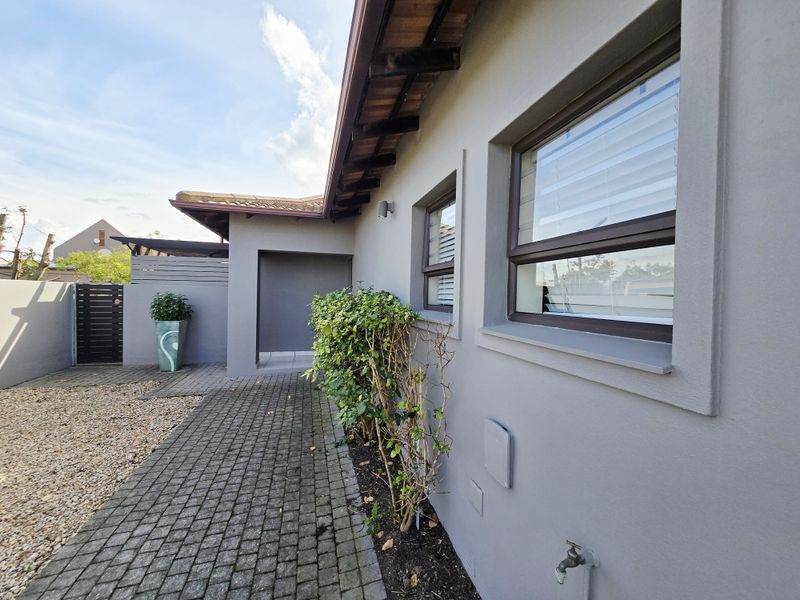 A well-maintained rear exterior view featuring a paved walkway, grey rendered walls, and aluminium window frames with a functional outdoor tap.