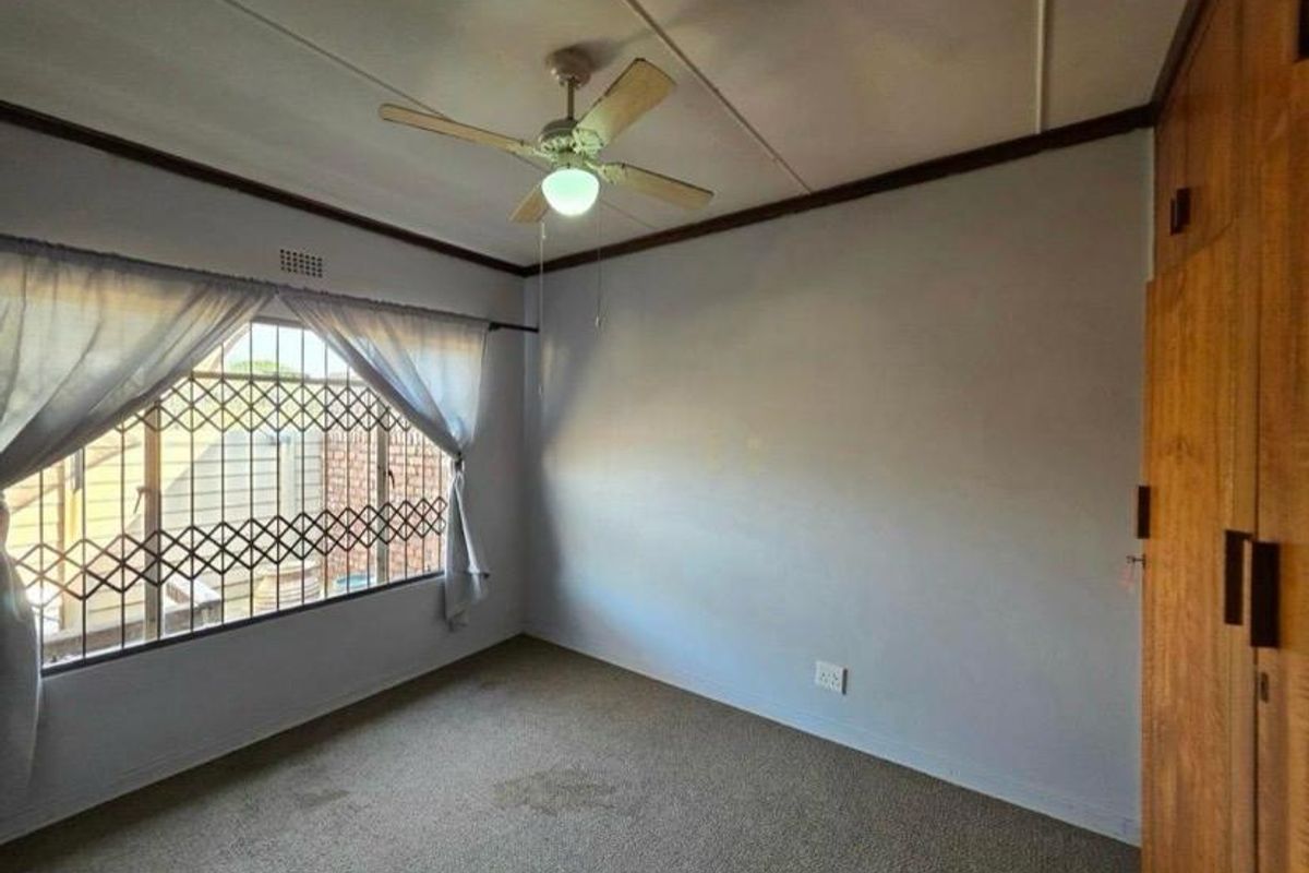 A spacious bedroom featuring built-in wooden cupboards, a ceiling fan, and large windows fitted with security gates.