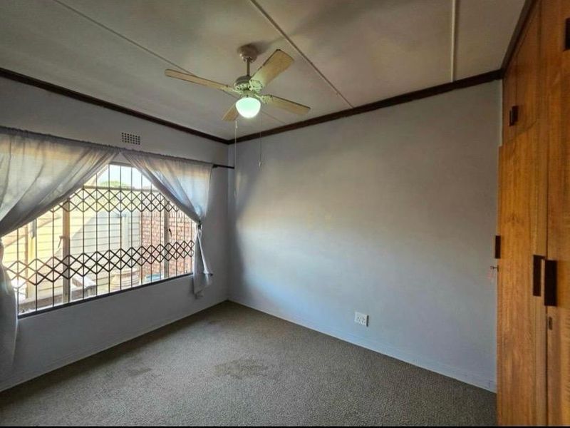 A spacious bedroom featuring built-in wooden cupboards, a ceiling fan, and large windows fitted with security gates.