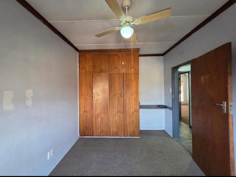 A vacant bedroom featuring built-in wooden cupboards, a built-in desk, and a ceiling fan, requiring cosmetic updates to the wall finishes.