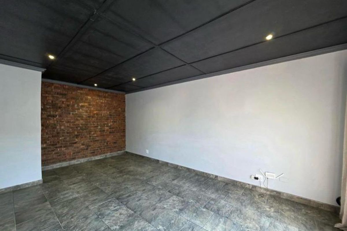 An empty living room featuring a striking exposed brick feature wall, dark painted ceiling with recessed lighting, and neutral tiled flooring.