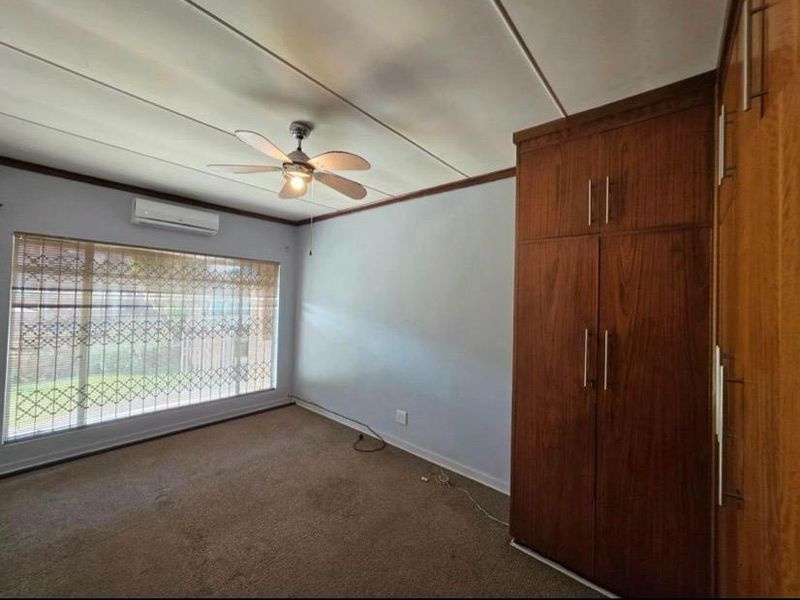 A spacious bedroom featuring built-in wooden cupboards, a ceiling fan, split-system air conditioning, and large windows with security bars.