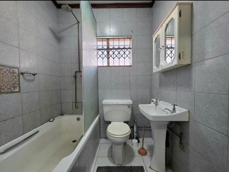 A functional bathroom featuring a pedestal basin, toilet, and a bathtub with a shower head and glass screen, finished with full-height wall tiling.
