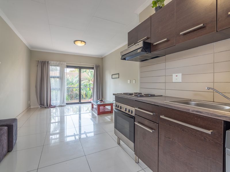 Open-plan living area featuring a modern kitchen with dark wood cabinetry, built-in oven, and tiled flooring, leading to a balcony with garden views.