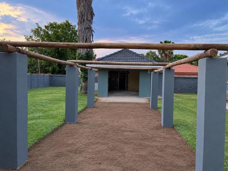 An outdoor patio area featuring a timber pergola structure supported by concrete pillars, overlooking a garden with a boundary wall.