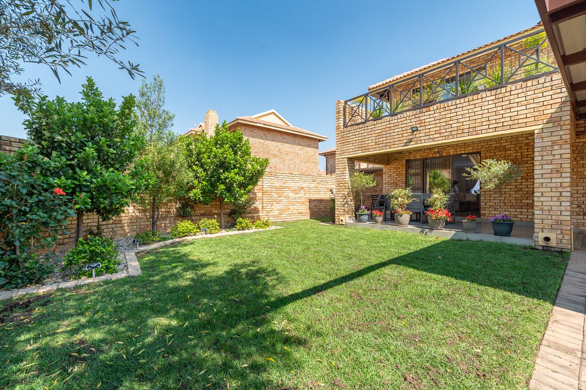 A well-maintained rear exterior view of a face brick townhouse featuring a covered patio, a private garden, and an upper-level balcony.