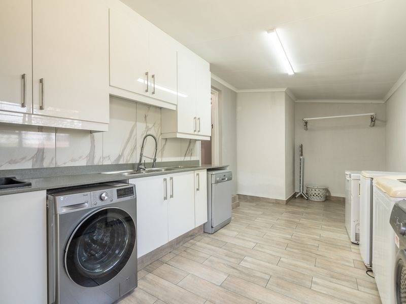 A clean and functional laundry room featuring white cabinetry, stone countertops, a stainless steel sink, and tiled flooring with ample space for appliances.