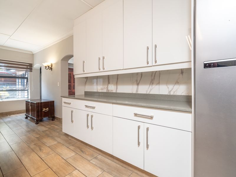 Modern kitchen featuring white cabinetry, stone countertops, marble-look splashback, and wood-look tile flooring, leading into an adjacent living area through an archway.