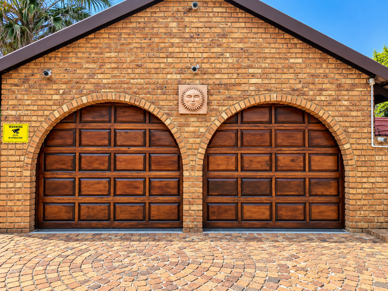 A well-maintained double garage featuring arched wooden doors, face-brick exterior, and a circular pattern paved driveway.