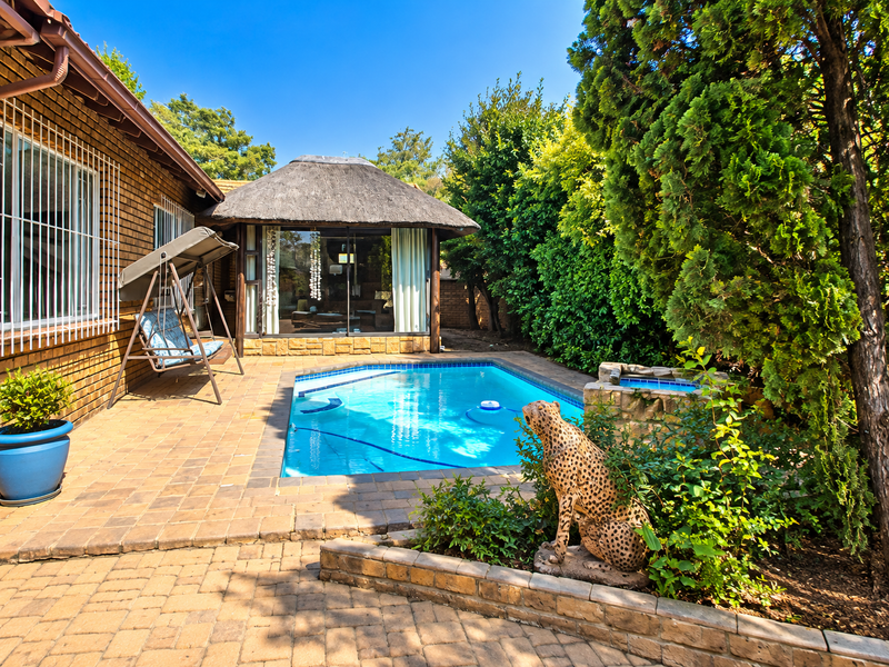 Outdoor pool area featuring a swimming pool, brick paved patio, and a thatched lapa structure set within a landscaped garden.