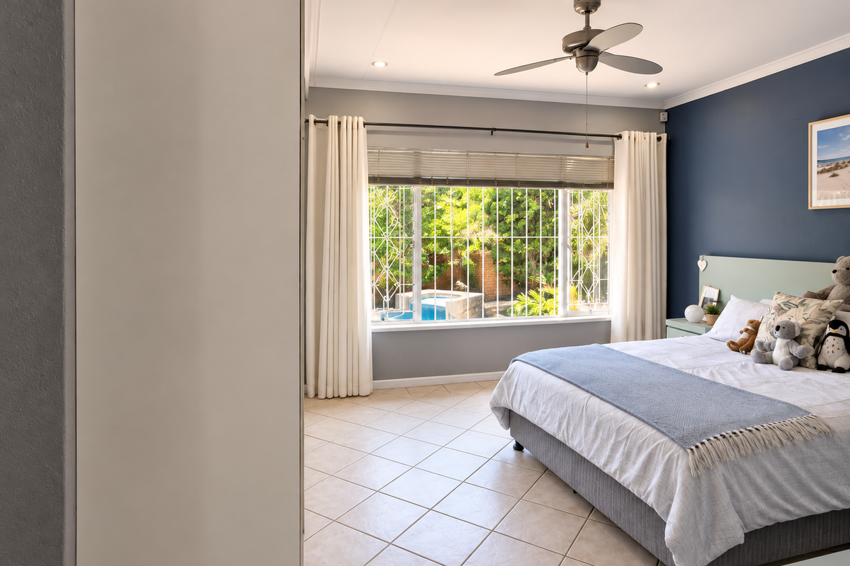 A well-maintained bedroom featuring tiled floors, a ceiling fan, and a large window with security bars overlooking the garden and pool area.