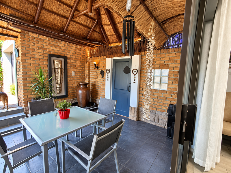 A covered outdoor patio featuring a rustic thatched roof, exposed brick walls, and dark tiled flooring, providing a seamless transition to the interior living space.