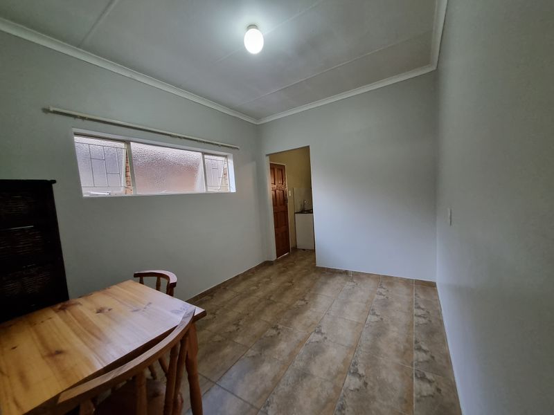A simple, neutrally painted living area featuring tiled flooring, a horizontal window, and direct access to an adjacent kitchen space.