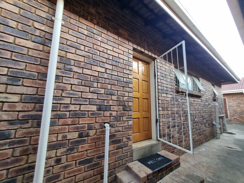 Rear exterior view of the cottage featuring face brick walls, a timber entrance door with a security gate, and concrete paving.
