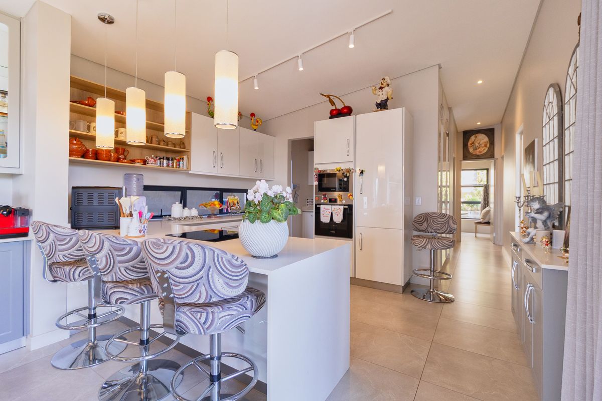 Modern kitchen featuring a central island with breakfast bar, built-in appliances, sleek cabinetry, and contemporary pendant lighting, leading into a bright hallway.