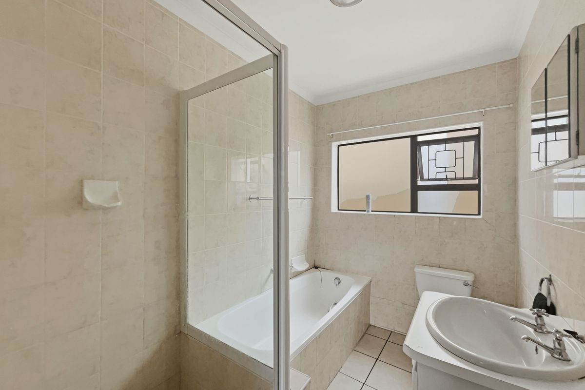 A functional bathroom featuring a built-in bathtub with a glass screen, a vanity basin, and a toilet, finished with neutral ceramic wall and floor tiles.