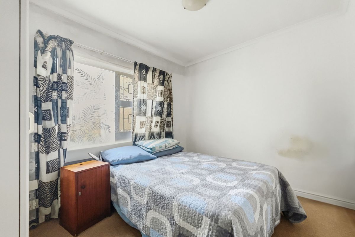 A bedroom featuring a built-in cupboard, window with security bars, and ceiling light fixture, showing signs of wall damage and wear.