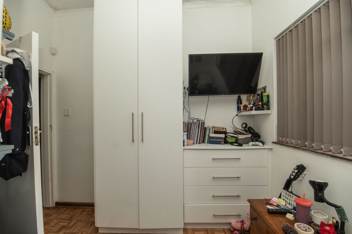 A bedroom featuring built-in white cabinetry, including a wardrobe and drawer unit, complemented by vertical blinds and parquet flooring.