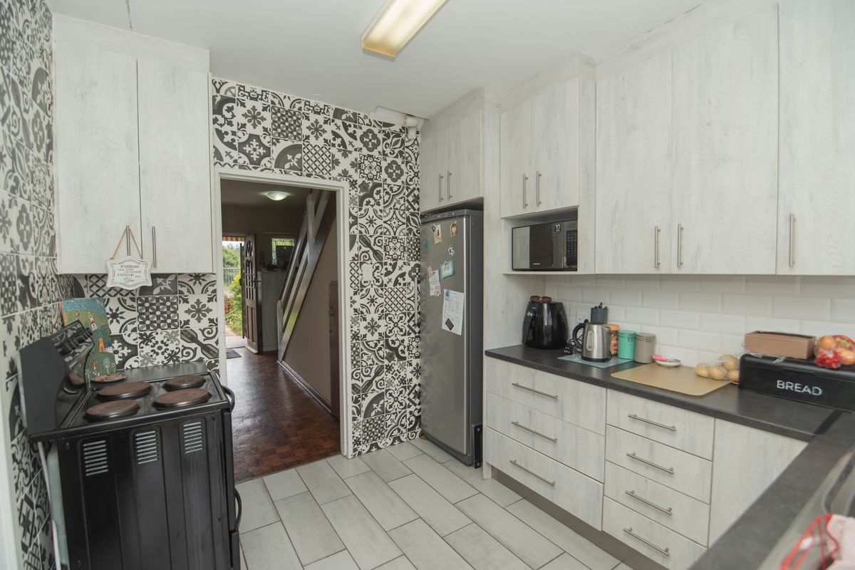 A functional kitchen featuring light wood-grain cabinetry, a striking patterned feature wall, subway tile backsplash, and ceramic floor tiles with direct access to the hallway.