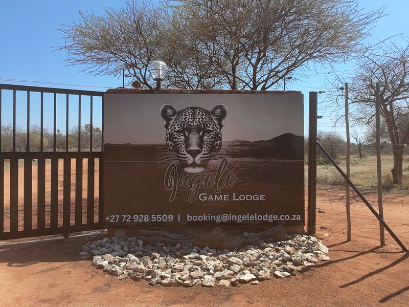 The entrance to the property features a branded signage board, metal security gate, and electric fencing, marking the approach to the game lodge.