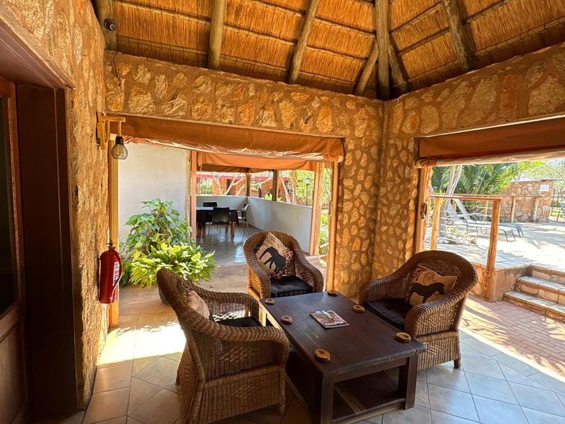 A rustic covered patio featuring a traditional thatched roof, exposed timber rafters, and stone-clad walls, opening onto an outdoor terrace area.