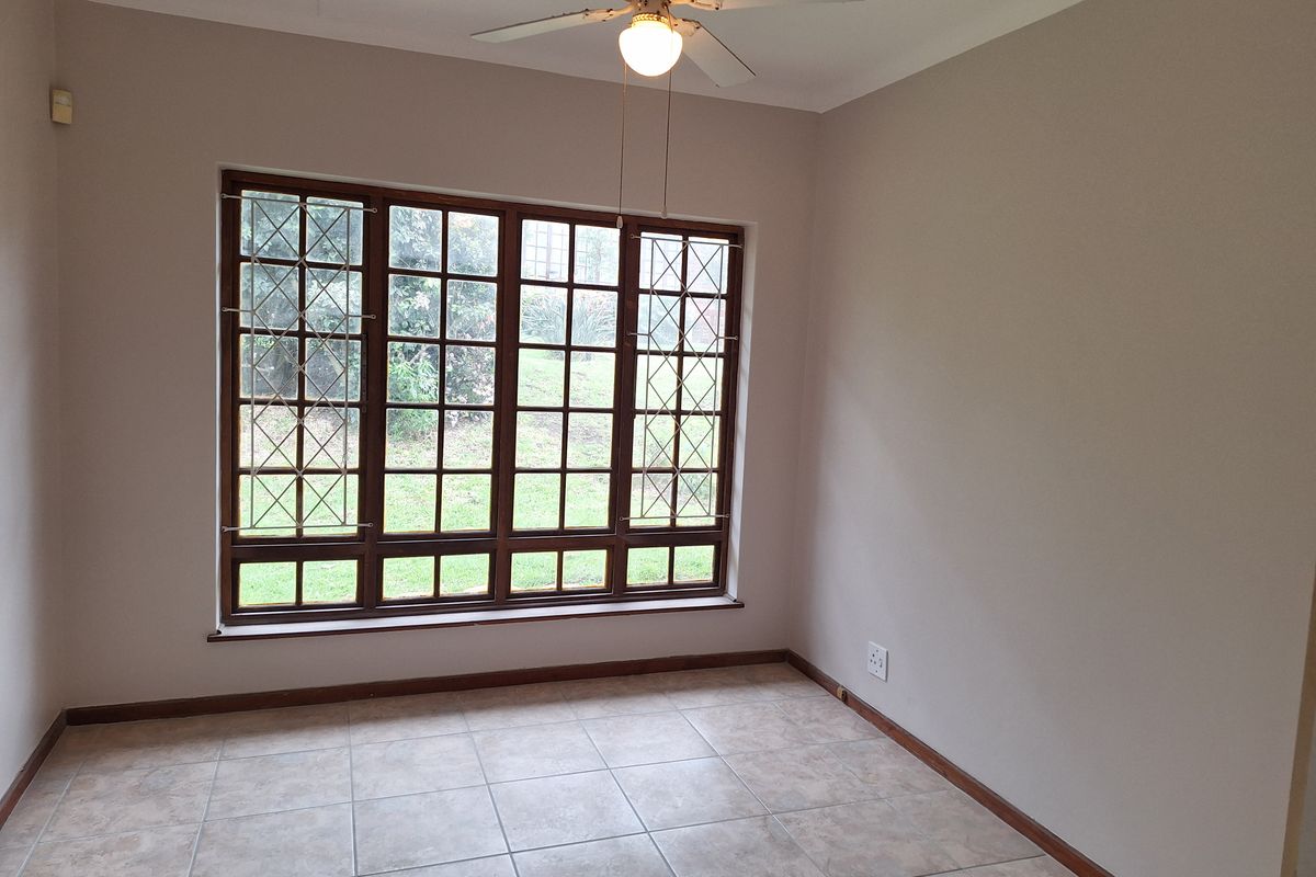 A bright, empty bedroom featuring neutral walls, tiled flooring, a ceiling fan, and large windows with security bars overlooking the garden.