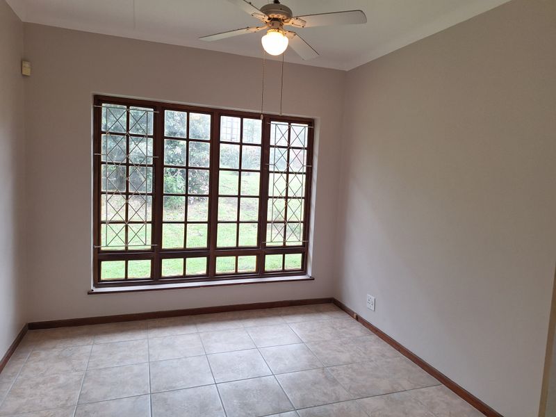 A bright, empty bedroom featuring neutral walls, tiled flooring, a ceiling fan, and large windows with security bars overlooking the garden.