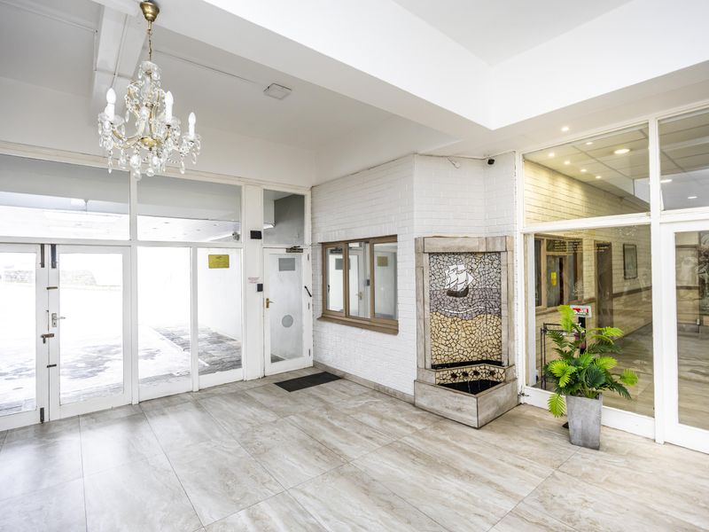 A bright and spacious apartment building entrance lobby featuring tiled flooring, glass panel doors, and a decorative mosaic water feature.