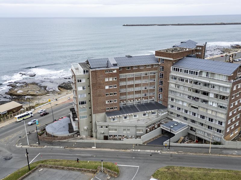 Aerial view of a multi-story apartment building featuring a brick facade and rooftop solar panels, situated along the coastline.