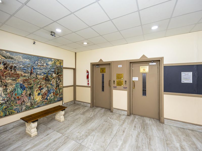 A well-maintained apartment building lobby featuring tiled flooring, a suspended ceiling with recessed lighting, and dual passenger lift access.