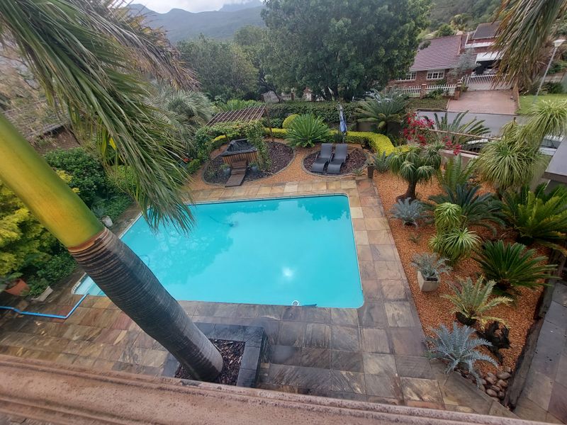 An elevated view of a well-maintained swimming pool area featuring stone paving, a wooden pergola, and established garden landscaping.