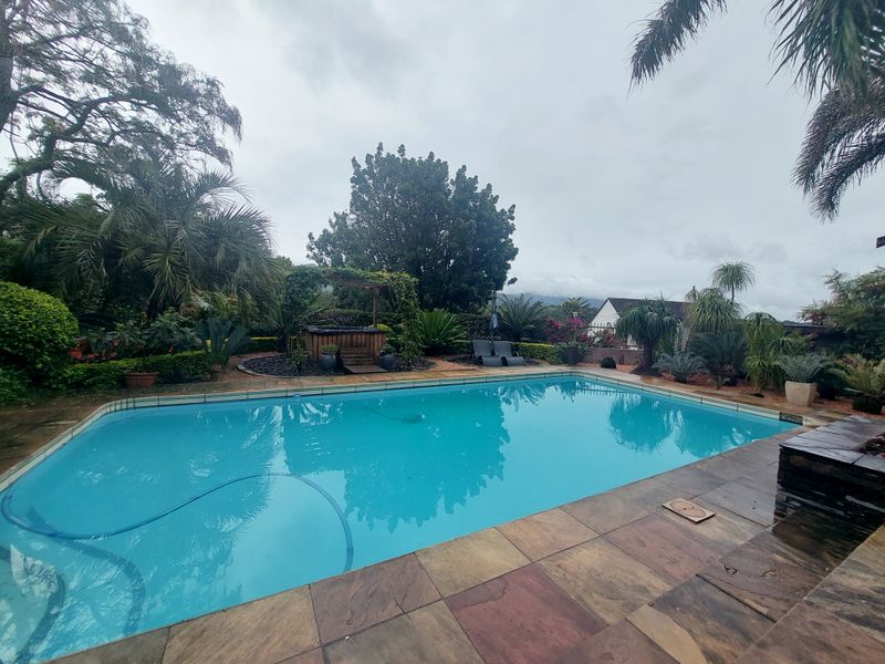 A well-maintained outdoor swimming pool area featuring stone decking, lush garden landscaping, and a wooden pergola structure.