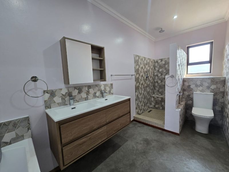 Modern bathroom featuring a double vanity with wood-finish cabinetry, a walk-in shower with decorative tiling, and a separate toilet area.