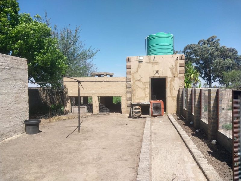 Rear exterior view of the small holding featuring a concrete walkway, brick boundary wall, and a water storage tank mounted on a masonry structure.