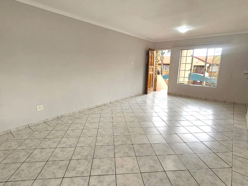 A spacious, empty living area featuring neutral wall finishes, tiled flooring, and natural light through a window with security bars.