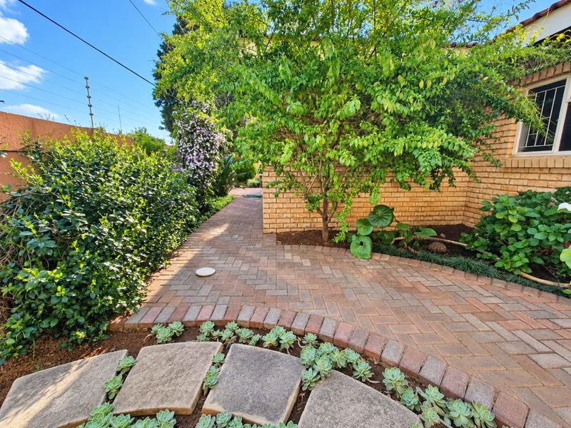 A well-maintained garden area featuring brick paved pathways, established trees, and face brick exterior walling with security bars on the window.