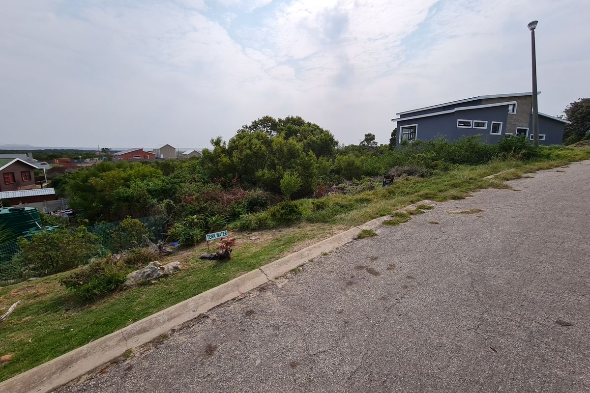A street-level view of a residential vacant land plot featuring an asphalt road, concrete curbing, and surrounding neighborhood vegetation.