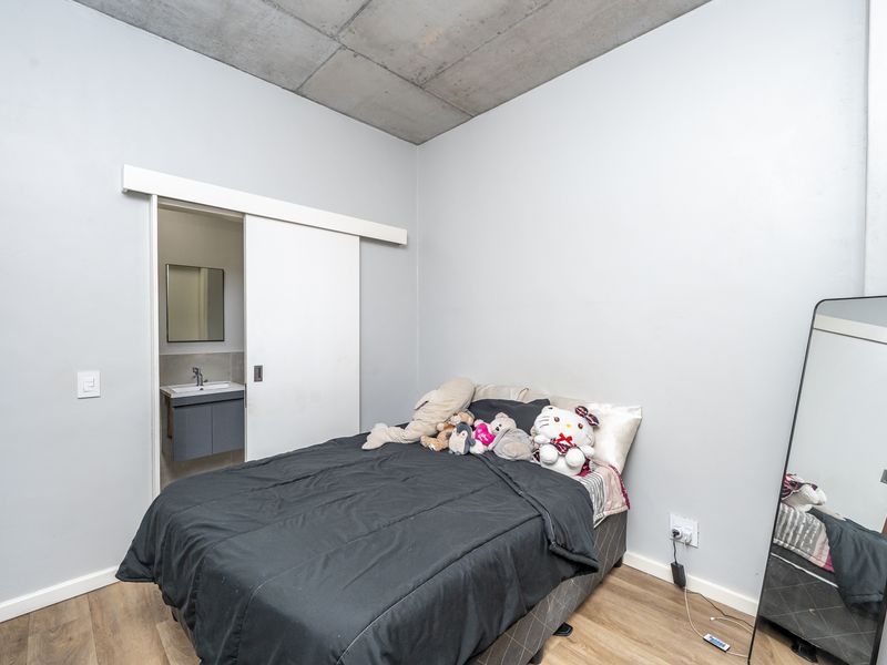 A modern bedroom featuring an exposed concrete ceiling, laminate flooring, and a convenient sliding door leading to an en-suite bathroom.